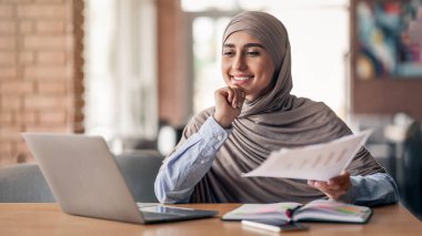 Kafede otururken, dizüstü bilgisayar kullanarak ve belgeleri kontrol ederek çalışan başarılı Müslüman kadın yönetici, boşluğu kopyala