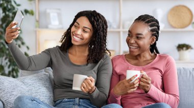 Joyful black women besties taking selfie on smartphone together while drinking tea at home, copy space. Cheerful african american young girlfriends having fun together on weekend, drinking coffee