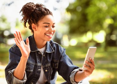 Cep telefonlu siyahi kadın dışarıda oturmuş el sallayarak video görüşmesi yapıyor. Uzaktan İletişim Konsepti. Seçici Odaklanma