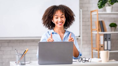 İngilizce öğrenmek, sağlık koruması ve mükemmel sonuçlar. Mutlu, Afro-Amerikan genç bir kadın başparmağını kaldırıyor ve sınıfın iç kısımlarındaki öğrencilere bakıyor.