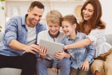 Ebeveynler ve çocuklar dijital tablet kullanıyorlar. Evdeki koltukta birlikte oturuyorlar. Aile Eğlencesi Konsepti