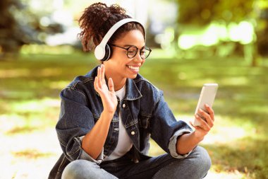 Video çağrısı. Cep telefonlu gülümseyen Afrikalı kadın dışarıda parkta oturan uzaktan kumandalı arkadaşla iletişim kuran akıllı telefona merhaba diyor. Modern Online İletişim Konsepti. Seçici Odaklanma