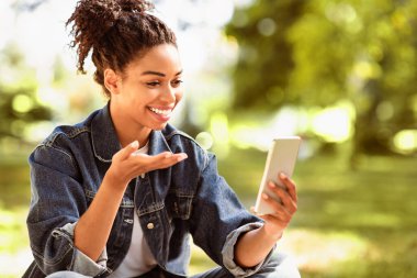 Telefonlu neşeli siyahi bayan parkta otururken video görüşmesi yapıyor. Öğrenci Kadın Cep Telefonu Kullanarak Online Video Konferansı Yapıyor. Modern Uzaktan İletişim Konsepti. Seçici Odaklanma