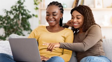 Kanepede oturup dizüstü bilgisayar kullanan iki mutlu siyah kadın. İnternet sörfü, boş alan. Gülümseyen Afrikalı Amerikalı kız arkadaşlar hafta sonunun keyfini çıkarıyorlar, sosyal medyayı birlikte kontrol ediyorlar.