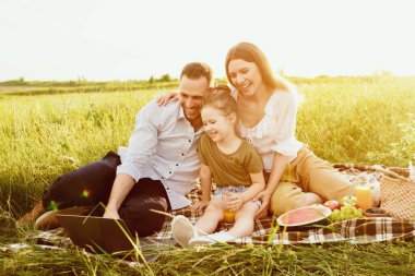 Birlikte eğleniyoruz. Güzel kızlarıyla gülümseyen ebeveynler piknik yapıyor ve bilgisayar kullanıyor.