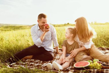 Eğleniyorum. Üç kişilik neşeli aile bir battaniyenin üzerinde dinleniyor, yiyeceklerle oynuyorlar, boş alanlar var.