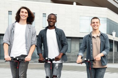 Arkadaşlık kavramı. E-scooter kullanan ve kameraya bakan sıradan, etnik çeşitlilikte üç genç.