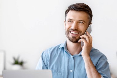 Ofiste dizüstü bilgisayarla çalışırken cep telefonuyla konuşan gülümseyen iş adamı, cep telefonu iletişiminin keyfini çıkaran, iş yerinde keyifli bir telefon konseri veren erkek girişimci,