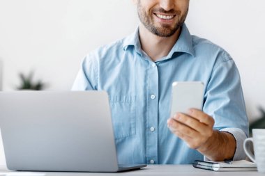 Yakışıklı erkek ofis yöneticisi iş yerinde akıllı telefon kullanıyor, masada oturuyor, sosyal medyayı kontrol ediyor veya uygulamayı tarıyor, mesaja cevap veriyor, ara veriyor, ekrana bakıyor ve gülümsüyor, boşluğu kopyalıyor.