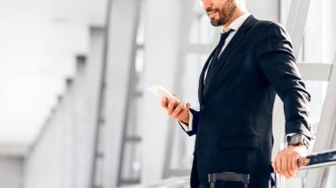 Yakışıklı sakallı iş adamı uçuş beklerken e-postayı kontrol ediyor, havaalanının penceresinin yanında duruyor, kopyalama alanı. Akıllı telefon kullanarak şık takım elbiseli orta yaşlı girişimci