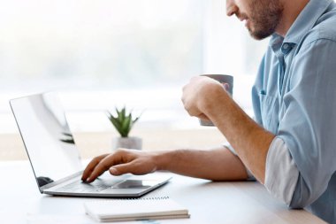 Yakışıklı erkek serbest çalışan, dizüstü bilgisayarda sosyal medyada gezinen ve sabah kahvesi içen, ev ofisinin penceresinin yanında oturan, uzaktan işten ve online işten hoşlanan, yan görünüm, uzay kopyası