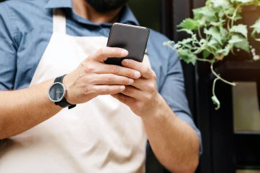 COVID-19 salgını sırasında yeni normal ve çiçek teslimatı hizmeti ve siparişler. Önlüklü genç erkek çiçek dükkanının yakınındaki telefonda dışarıda saksı ve vazolarda bitkilerle mesaj yazıyor, panorama.