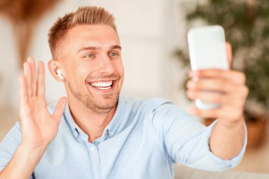Çevrimiçi Video Çağrısı. Sanal konferans yapan gülümseyen adamın portresi, cep telefonu tutan, kablosuz kulaklık takan, web kamerasına el sallayan. İletişim Konsepti. Akıllı telefon kullanan bir adam sohbet ediyor.