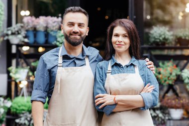 Shop and flower studio owners, waiters in eco cafe and greeting customers outdoor. Smiling young attractive man and woman in aprons, on flower studio background with decor elements and plants