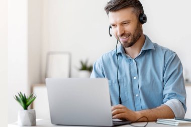 Çevrimiçi kulaklıklı yakışıklı genç adam, dizüstü bilgisayarda Webinar izliyor, uzaktan eğitimin tadını çıkarıyor ve internetten öğreniyor, evde masa başında oturuyor, bilgisayar ekranına bakıyor, boş alana bakıyor.