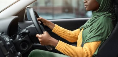 Young Driver. Serious black muslim lady in hijab driving car, holding steering wheel and looking ahead focused on the road, young african woman in headscarf riding medern automobile in city, side view
