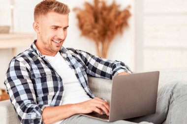 Serbest ve Uzaktan Çalışma Konsepti. Kareli tişörtlü olumlu bir adamın portresi internette dizüstü bilgisayar kullanarak sohbet ediyor, rahat oturma odasında kanepede oturuyor. Gülümseyen serbest yazar evde bilgisayarla çalışıyor.