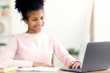 Afro-Amerikalı ergen kız laptopta ödevini yapıyor. Kapalı bir masada oturuyor. Seçici Odaklanma