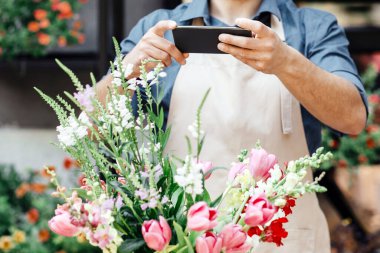 Çiçekçi, teslimat, düzen ve modern online satış. Bin yıllık meşgul erkek sahibi önlüklü ve koruyucu maskeli şık bir buket yapıyor ve fotoğraf çekiyor, stüdyoda, boş alanda, açık havada.