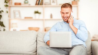 Müşteri Destek Konsepti. Cep telefonuyla konuşan, bilgisayarla çalışan oturma odasındaki kanepede oturan gülümseyen adamın portresi. Telekomünikasyon ve telesekreter. Dizüstü bilgisayar kullanan kendine güvenen yetişkin