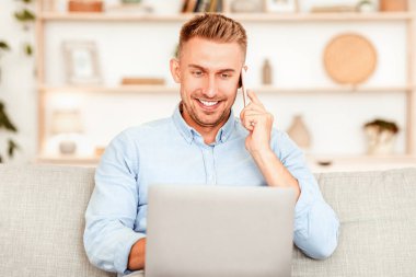 Kurumsal İletişim. Gülümseyen genç girişimcinin portresi telefon görüşmesi yapıyor, dizüstü bilgisayarda yazıyor, oturma odasındaki kanepede oturuyor. İş için bilgisayar kullanan kendine güvenen yetişkin, İnternet 'te istemciye danışın