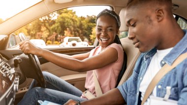 Genç siyahi erkek eğitmen, mutlu bayan öğrenciyi muayene ediyor, çizelgeye not alıyor, neşeli Afro-Amerikan bir kadının yanında oturuyor, araba sürüyor, yan görüş, kopyalama alanı, güneş ışığı...