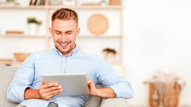 Gülümseyen adam dijital tablet kullanıyor modern dairesindeki koltukta oturuyor, boşluğu kopyalıyor.