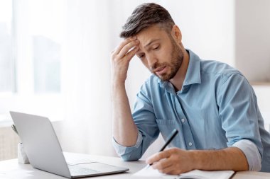 Ofiste dizüstü bilgisayarla çalışırken not alan, iş takvimi hazırlayan, son teslim tarihi stresli ve iş yerinde sorunları olan, endişeli yüz ifadesiyle alınına dokunan iş adamı.