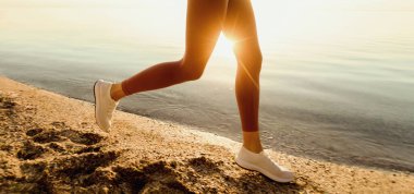 Sahilde koşan, dışarıda spor yapan, arka planda doğal olarak ılık gün doğumu yapan bir kadının görüntüsü.