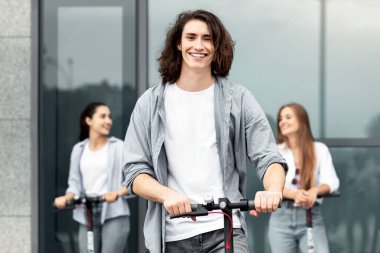 Gençlik Konsepti. Uzun saçlı neşeli adam stand-up scooter kullanıyor, arkadaşlarıyla vakit geçiriyor.