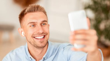 Çevrimiçi İletişim. Mutlu adamın video araması yaptığı, akıllı telefon tuttuğu, kablosuz kulaklık taktığı yakın plan portresi. Gülümseyen adam blog kaydediyor, selfie çekiyor, sosyal medya için video bloğu çekiyor.