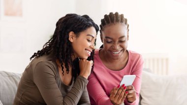 Gülümseyen Afro-Amerikan kadınları akıllı telefondan fotoğraf izleyip evdeki kanepede oturuyorlar. Cep telefonu kullanan iki neşeli zenci kız arkadaş, web sörfü ya da online alışveriş, boş alan