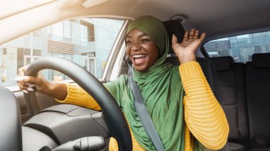 Yol Eğlencesi. Neşeli Afrikalı Müslüman kadın, arabada dans ediyor, yeni aracını kullanırken şarkı söylüyor, neşe saçan siyah kadın başörtüsü takıyor, şehirde yol yolculuğunun tadını çıkarıyor, düşük açılı manzara