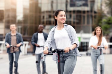 Hafta sonu. Genç ve güzel bir kız yazın sokakta elektrikli scooter kullanıyor, arkadaşlarıyla eğleniyor.