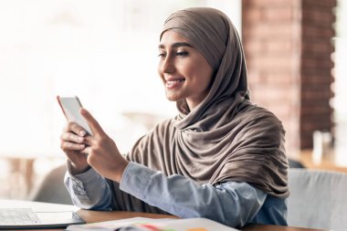Laptop kullanan genç Müslüman kadın kafede akıllı telefon kullanıyor, online iş yapıyor, boş yer var.