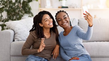 Cute black girlfriends sitting on floor by couch in living room and taking selfie together, using mobile phone, copy space. Happy african american women enjoying weekend at home