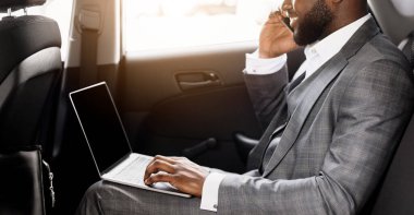 Genç siyahi iş adamı, ofise giderken arabada cep telefonu ve laptop kullanıyor. Diz üstü bilgisayarda çalışan Afrikalı Amerikalı girişimci ve iş toplantısına giderken akıllı telefondan sohbet eden