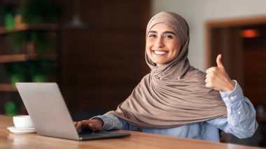Başparmak gösteren tesettürlü mutlu Müslüman kadın, kafede çalışıyor, dizüstü bilgisayar kullanıyor, kahve içiyor, fotokopi çekiyor. Yan bakışta gülümseyen Arap kadın dizüstü bilgisayarda yazı yazıyor, uzak işinden zevk alıyor.