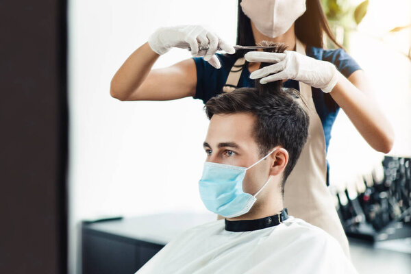 Young hairdresser in protective mask cutting hair of male client, close up