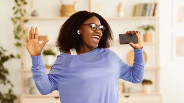 Heyecanlı zenci kızın evdeki akıllı telefonuna şarkı söyleyip kablosuz kulaklıkla müzik dinlediği bir fotoğraf.