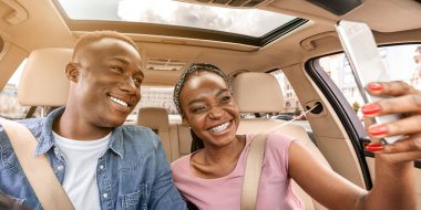Şirin, neşeli, siyah bir çift araba kullanıyor ve akıllı telefondan selfie çekiyor. Mutlu Afro-Amerikan milenyumu erkek ve kadın rahat bir arabayla tatile çıkarken cep telefonuyla fotoğraf çekiliyor.