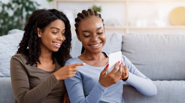 Neşeli siyahi kız arkadaşlar cep telefonundan fotoğraf çekiyorlar, evde vakit geçiriyorlar, fotokopi çekiyorlar. İki Afro-Amerikalı bayan oturma odasında kanepenin yanında oturuyorlar, akıllı telefon kullanıyorlar.