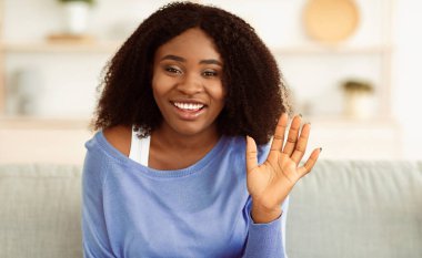 Selamlama Kavramı. Gülümseyen güzel Afro-Amerikan kadın portresi. El sallayarak merhaba diyor, kameraya bakıyor, evdeki koltukta oturuyor. Siyahi kadın merhaba diyor, gülüyor, geçmişi bulanık.
