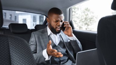 Öfkeli Afrikalı Amerikalı iş adamı cep telefonuyla konuşuyor ve iş toplantısına arabayla giderken laptop ekranına bakıyor, genç siyah yönetici arabanın arka koltuğunda oturuyor, çalışıyor, yer kopyalıyor.