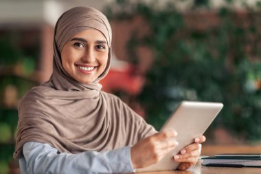 Kafede çalışan, dijital tablet kullanan, tesettürlü, mutlu bir Arap kadın. Başörtülü gülümseyen Müslüman kadın dijital ped tutuyor, kafeteryada kahve içerken blog yazıyor, internette çalışıyor.