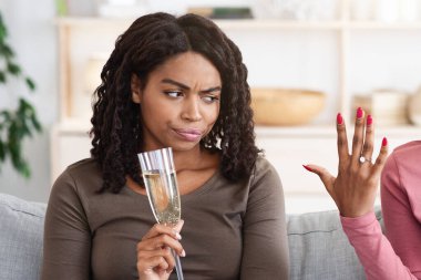 Happy black woman showing her upset and jealous girlfriend engagement ring. Annoyed african american lady cannot share her female friend happiness, drinking champagne together at home