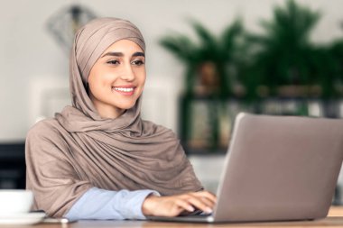 Kafe 'de çalışan neşeli Müslüman kız dizüstü bilgisayar kullanıyor ve kahve içiyor.