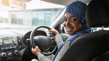 Otomobil almak. tesettürde gülümseyen Müslüman kadın yeni arabayı test ediyor, ön koltukta oturuyor, kamerada yarım yamalak dönmüş, direksiyonu tutuyor, modern otomobilin keyfini çıkarıyor, bedava uzay aracı