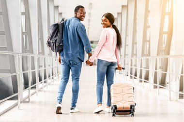 Çift Seyahat Konsepti. Romantik siyah eşler havalimanında bavullarıyla yürürken mutlu Afrikalı erkek ve kadınlar balayı tatiline gider, el ele tutuşur, kameraya bakar ve gülümserler.