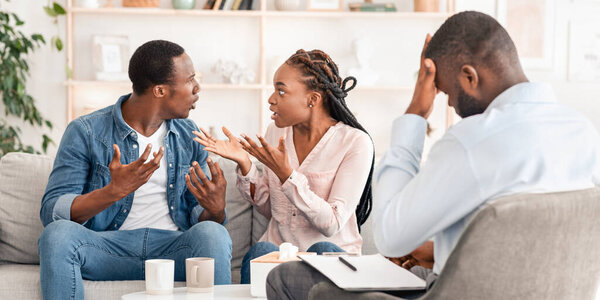 Брачный кризис. Молодые чернокожие супруги спорят на сеансе семейной терапии, сидят на диване в офисе психолога, свободное место
